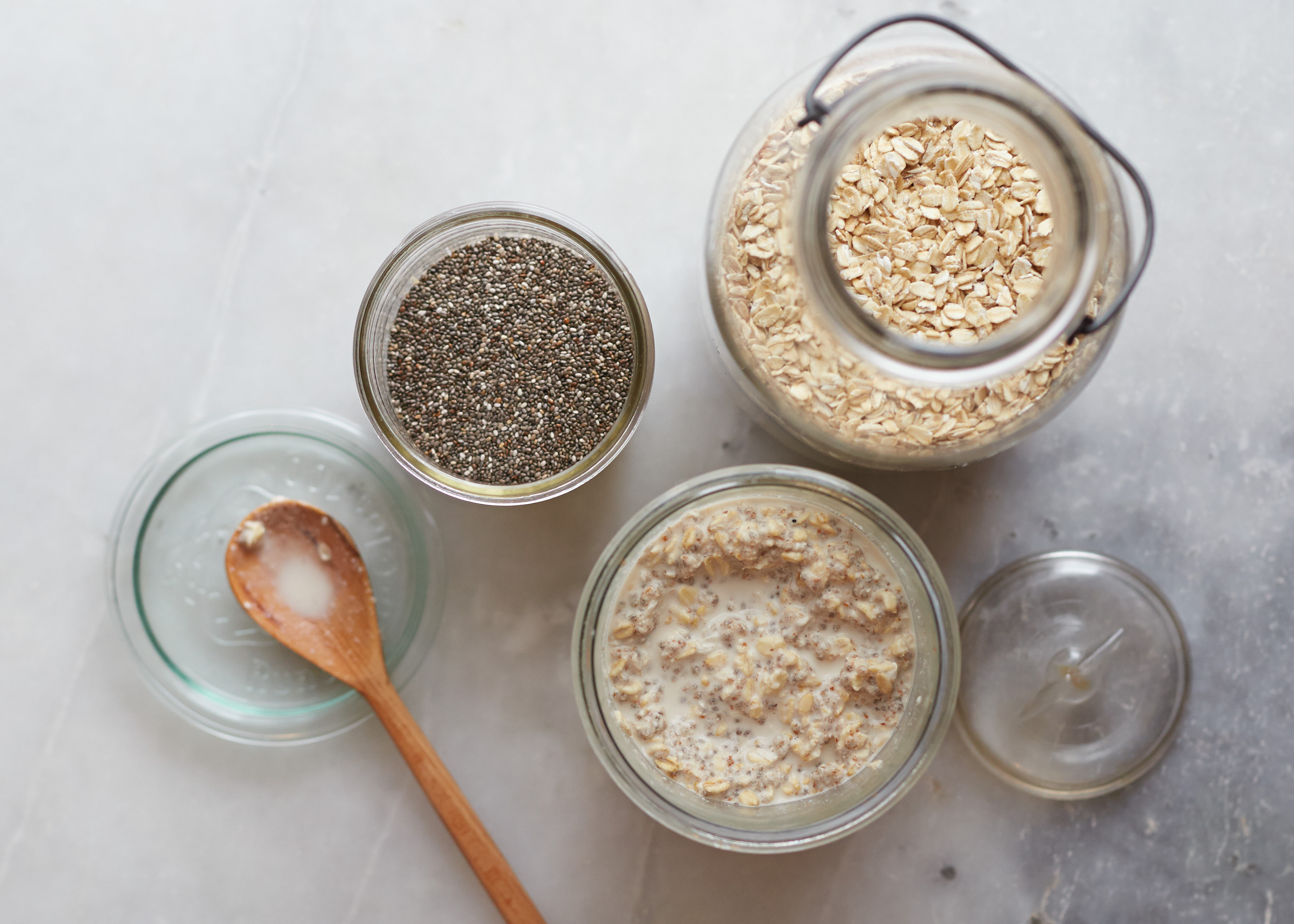 Rolled oats and chia seeds measured in small bowls next to a jar and spoon on a kitchen counter