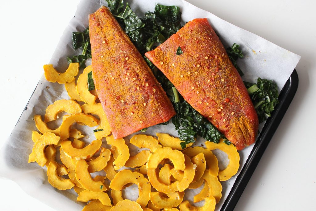 Salmon fillets on a sheet pan being seasoned with turmeric and spices next to lemon wedges on a kitchen counter