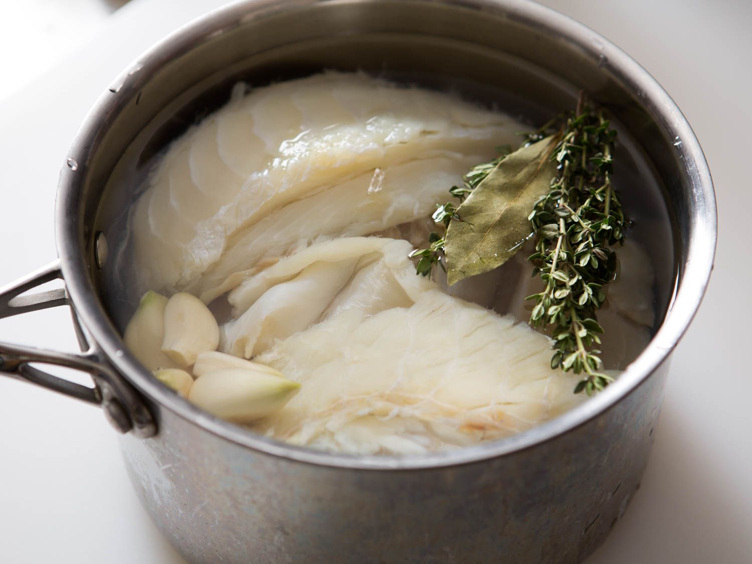 Salt cod pieces gently simmering in a small saucepan on a stovetop with light steam rising