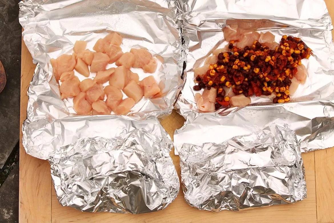 Seasoned chicken strips with sliced peppers and onions portioned onto foil sheets on a picnic table, ready to be folded into packets