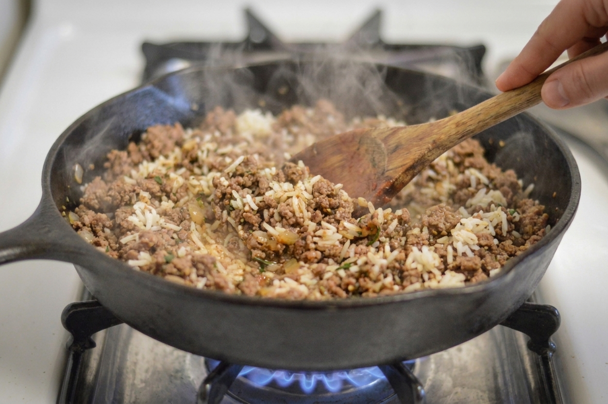 Seasoned ground beef and rice filling being stirred in a large skillet on a stove with steam rising