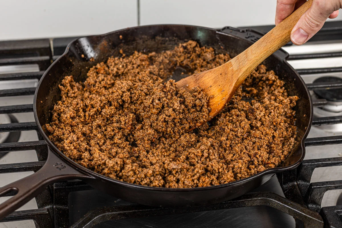 Seasoned ground beef cooking in a skillet with onions and spices, being stirred with a wooden spoon