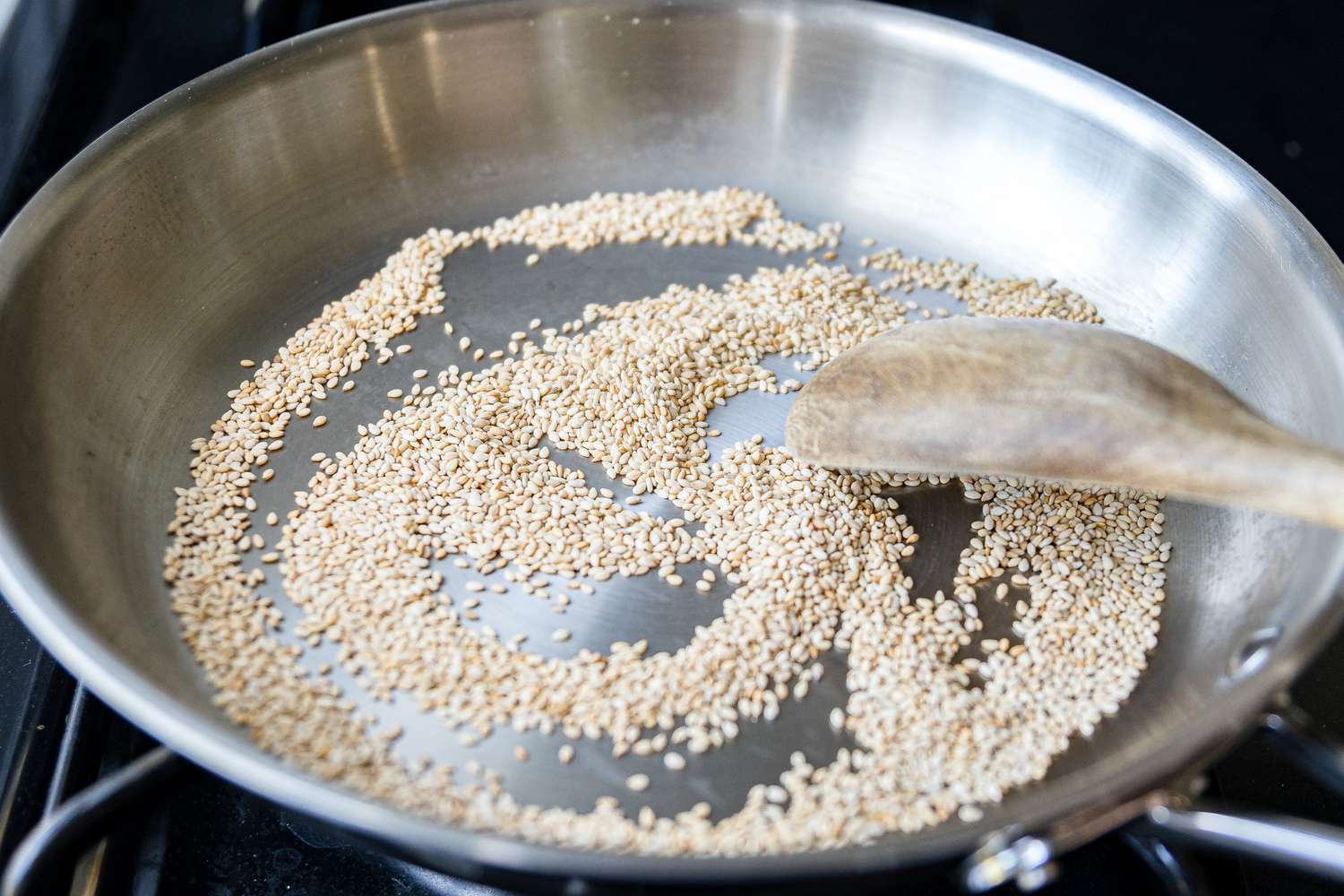 Sesame seeds being toasted in a small skillet on a stovetop with a wooden spoon stirring
