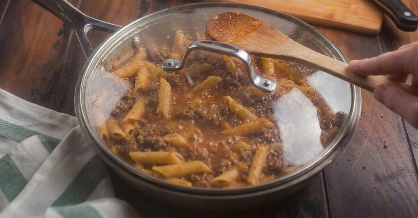 Short pasta simmering in a saucy meat mixture in a covered skillet on the stove