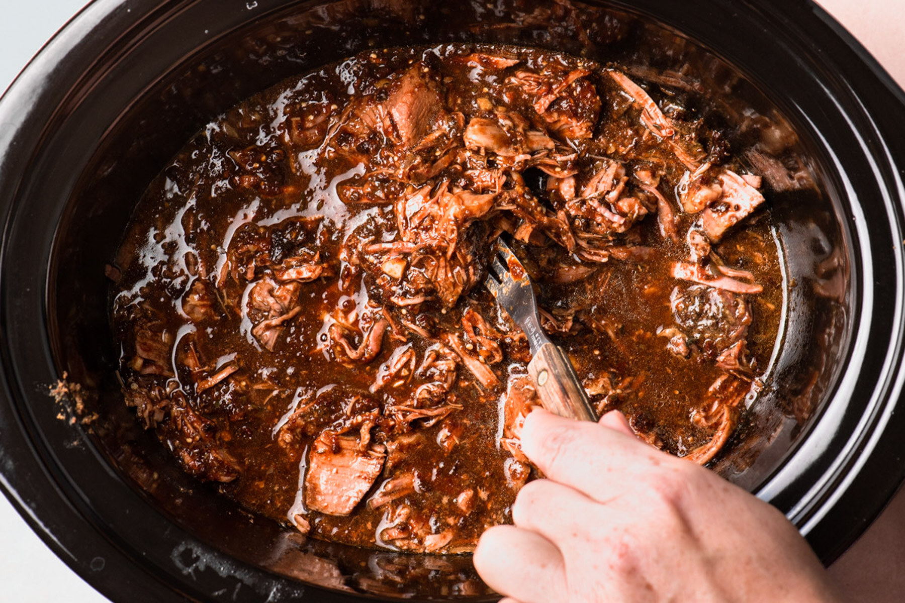 Shredded barbacoa in a glass meal prep container with cooking juices, set on a clean kitchen counter next to a lid