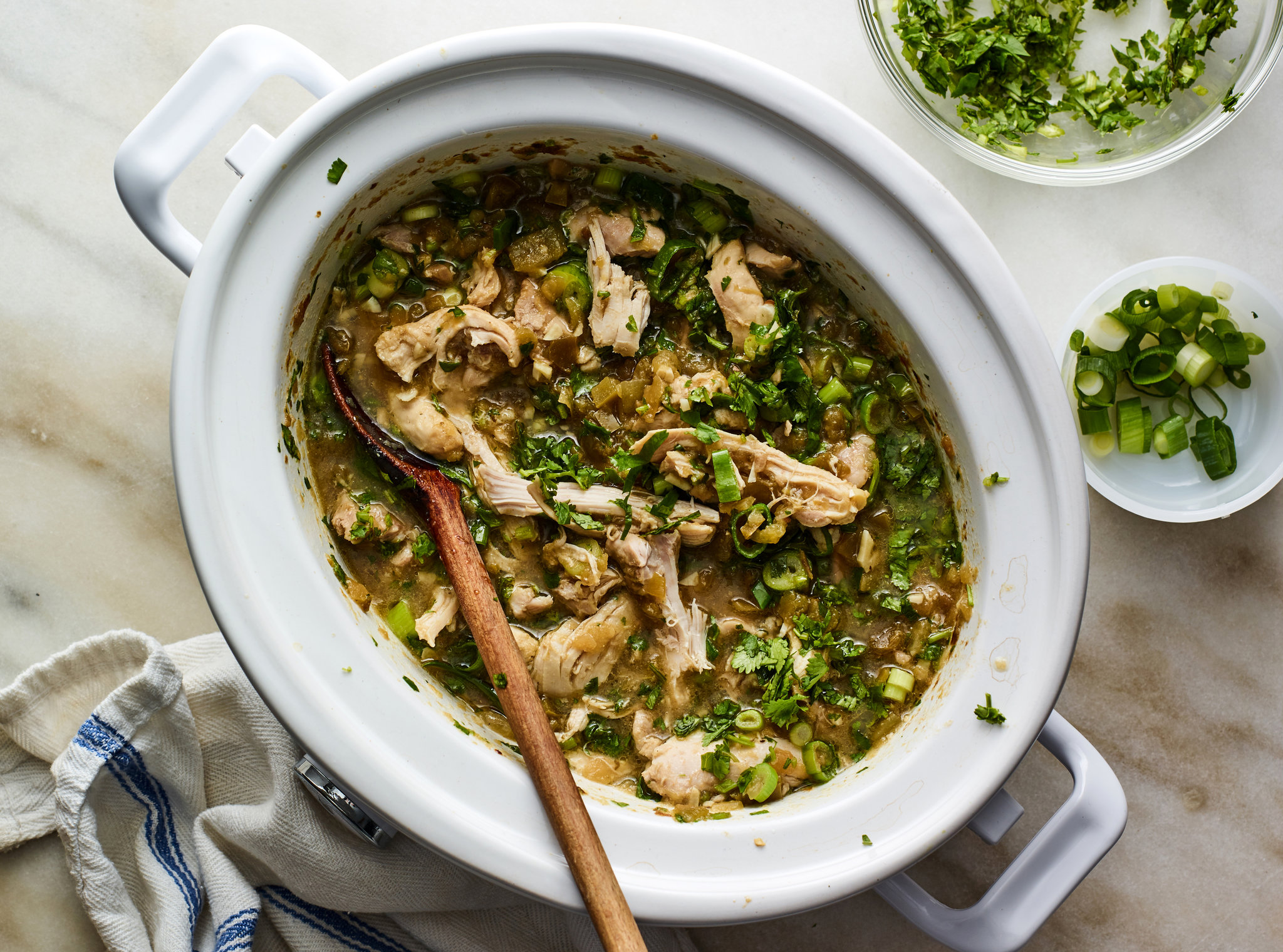 Shredded chicken coated in green salsa inside a slow cooker with a pair of tongs resting on the rim