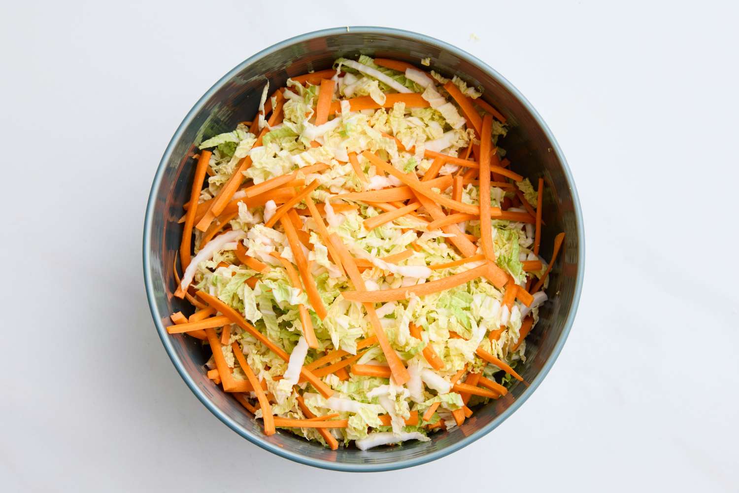 Shredded green and purple cabbage, carrots, and scallions in a large white bowl on a bright kitchen counter, ready to be tossed