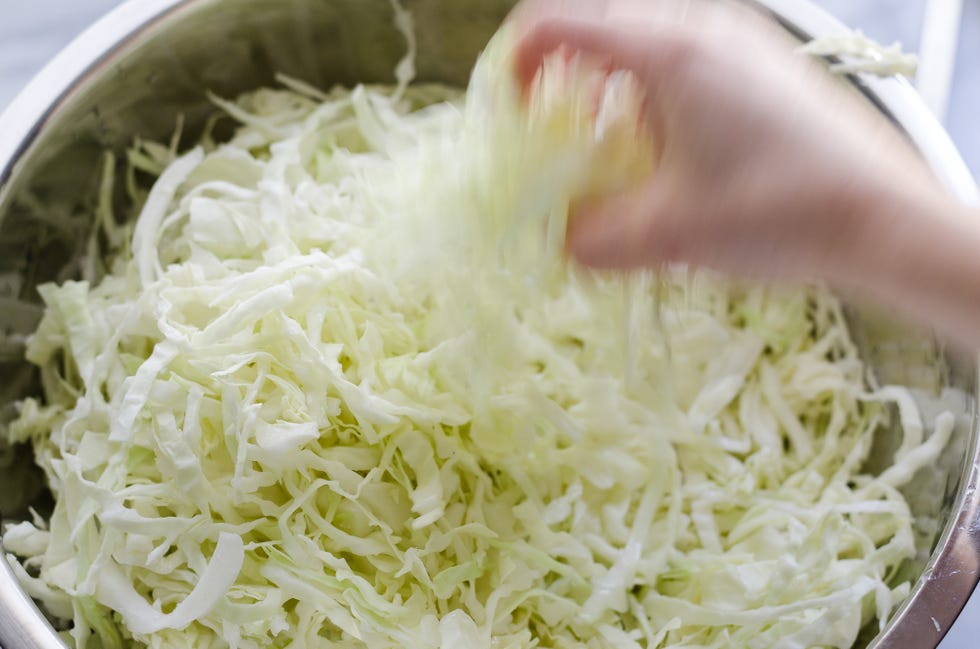 Shredded green and purple cabbage in a large mixing bowl with salt being sprinkled over the top on a kitchen counter