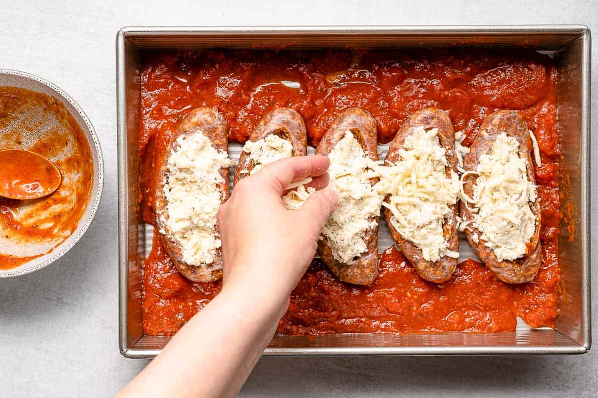 Shredded mozzarella in a bowl beside browned Italian sausage in a skillet and a can of crushed tomatoes on a kitchen counter