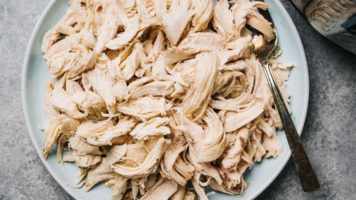 Shredded rotisserie chicken in a bowl next to a large skillet on a stove, ready to be reheated