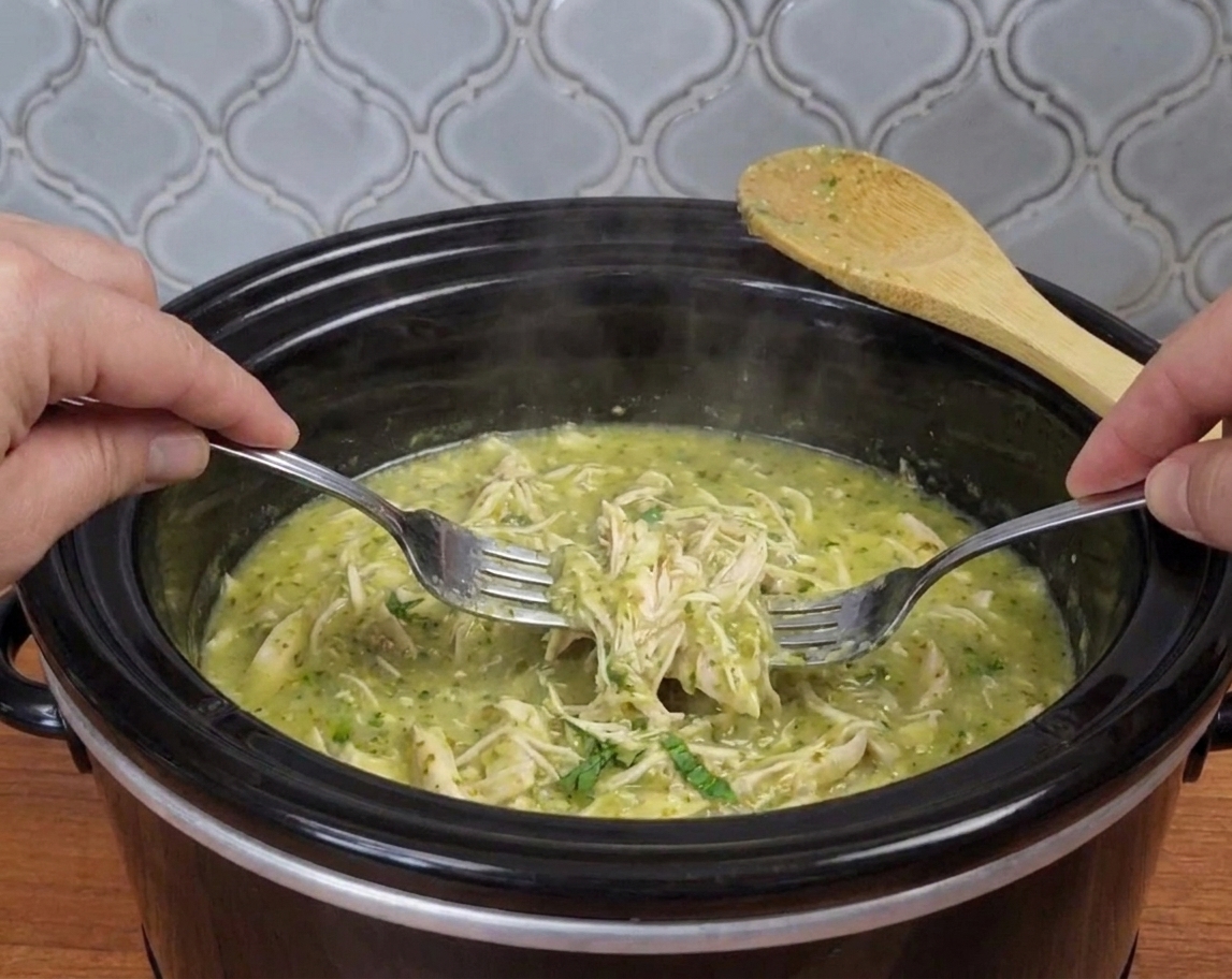 Shredded salsa verde chicken being pulled apart with two forks inside a slow cooker