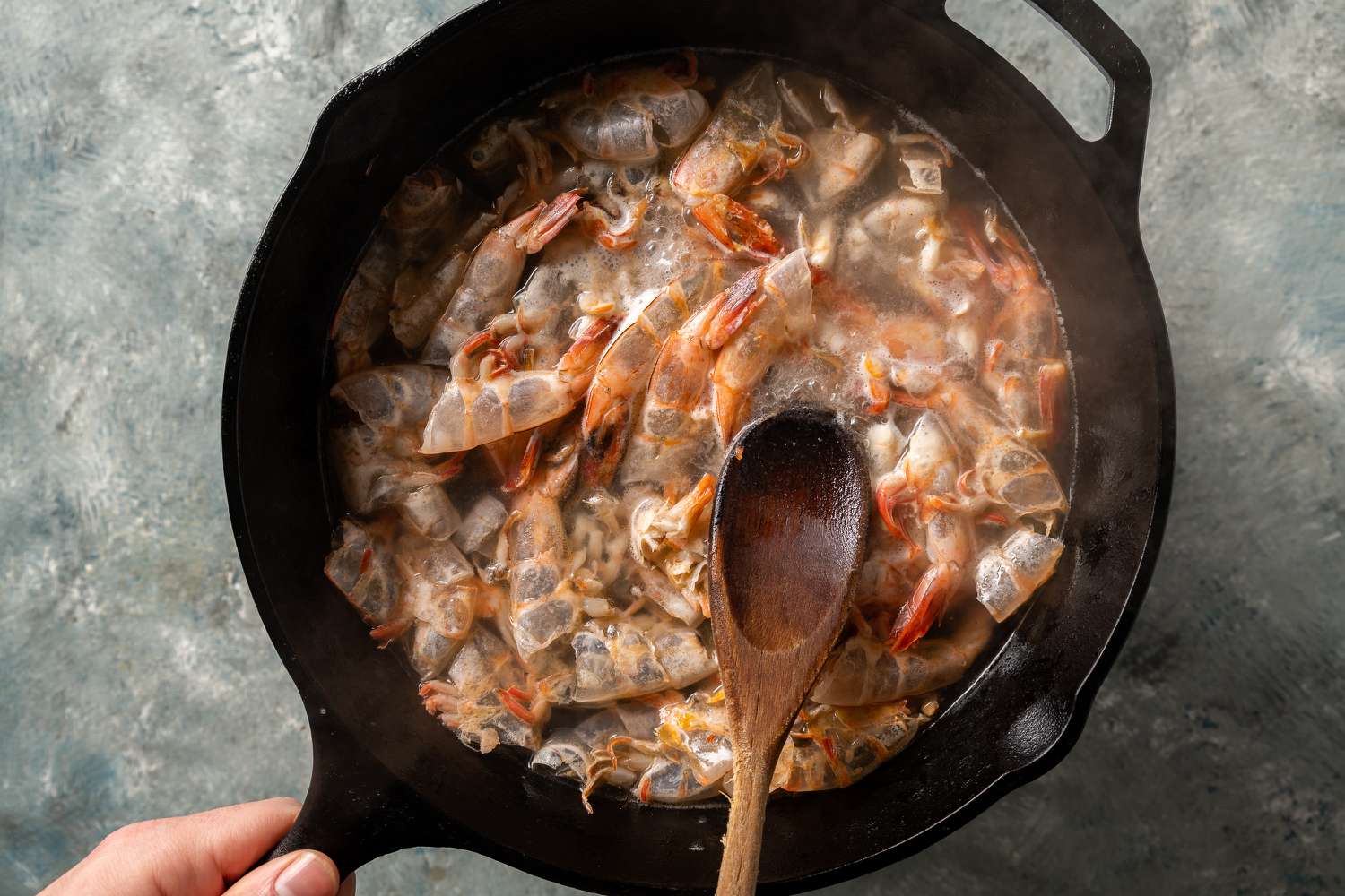 Shrimp shells and aromatics simmering in a pot of stock on a stovetop, with a wooden spoon resting nearby