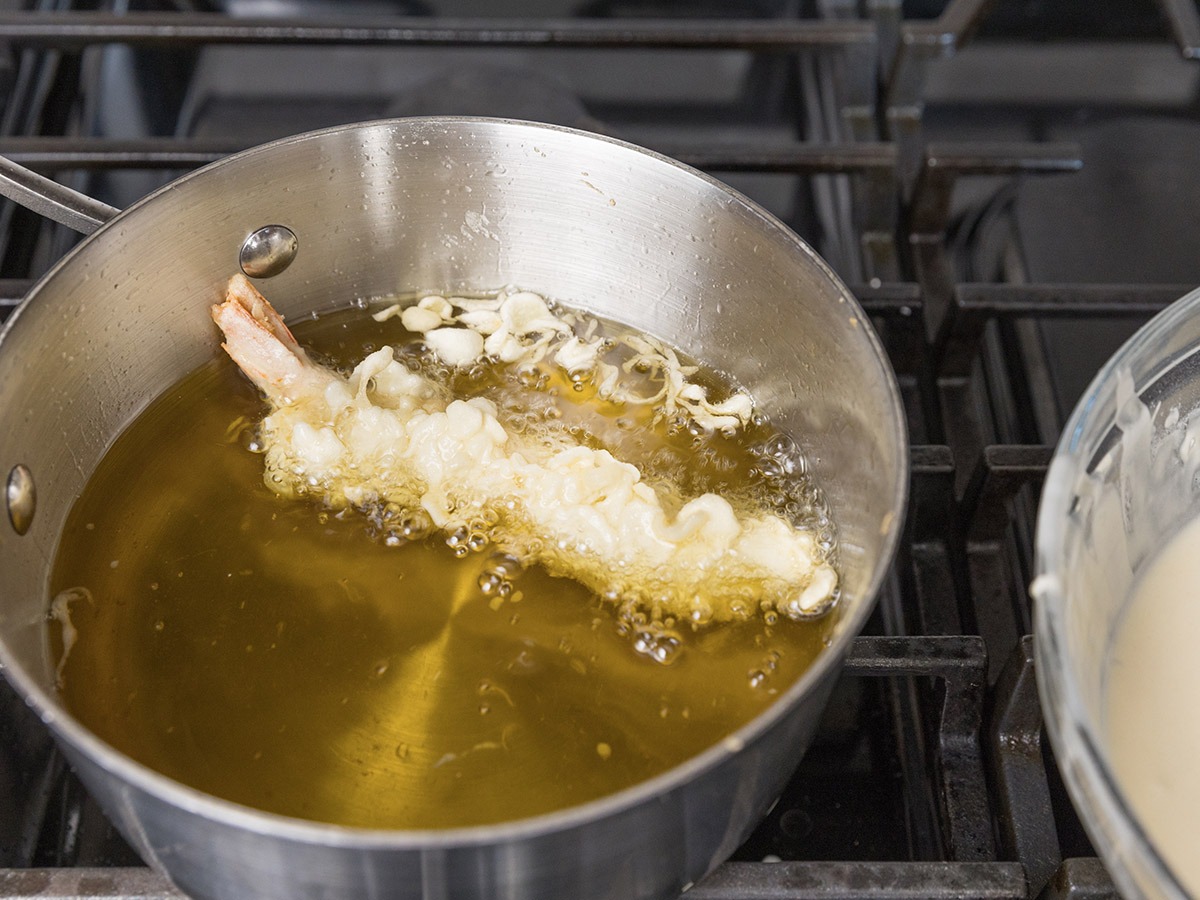 Shrimp tempura pieces bubbling in hot oil in a deep pot while a spider strainer lifts one piece