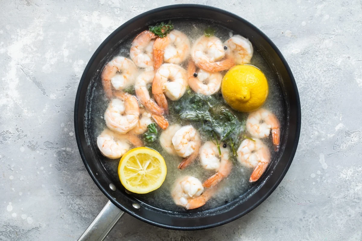 Shrimp turning pink in hot water in a stainless steel saucepan on a home stove, with a slotted spoon resting nearby