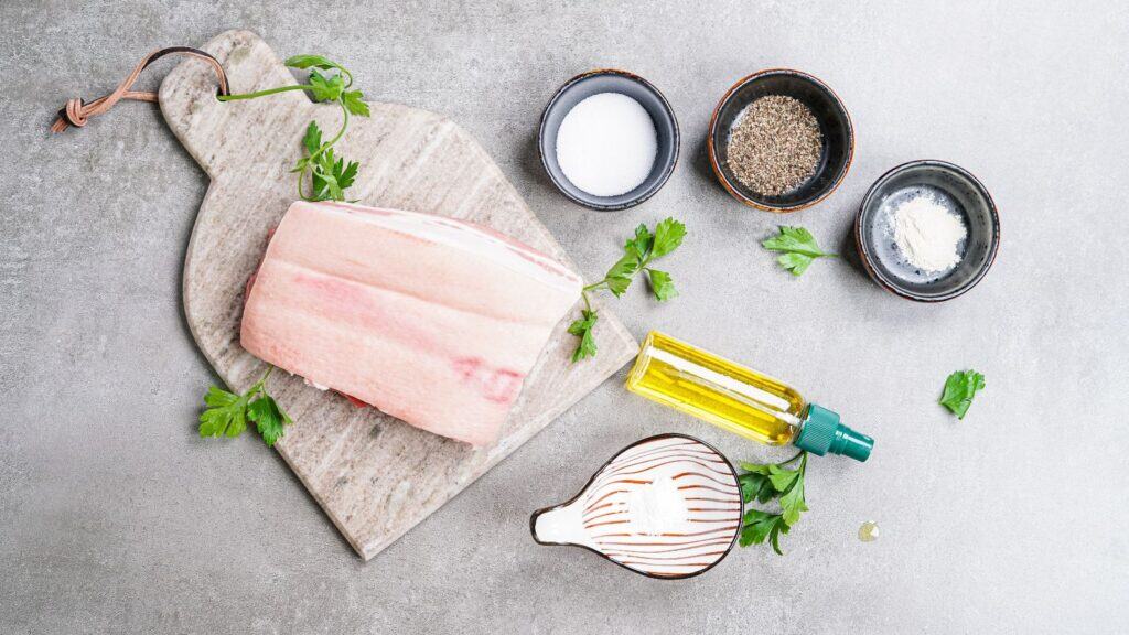 Skin-on pork belly on a cutting board surrounded by small bowls of kosher salt, baking powder, garlic powder, and smoked paprika on a kitchen counter