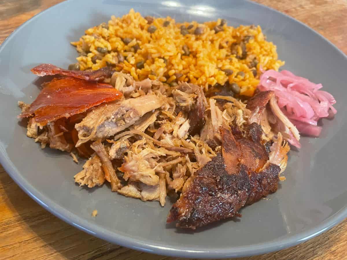 Sliced Puerto Rican pernil on a large platter with crisp golden skin, visible garlic and herb bits, and citrus wedges on a warm-toned wooden table