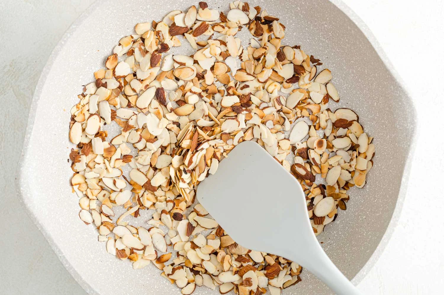 Sliced almonds toasting in a small skillet on a stovetop with a wooden spoon nearby in a warm home kitchen