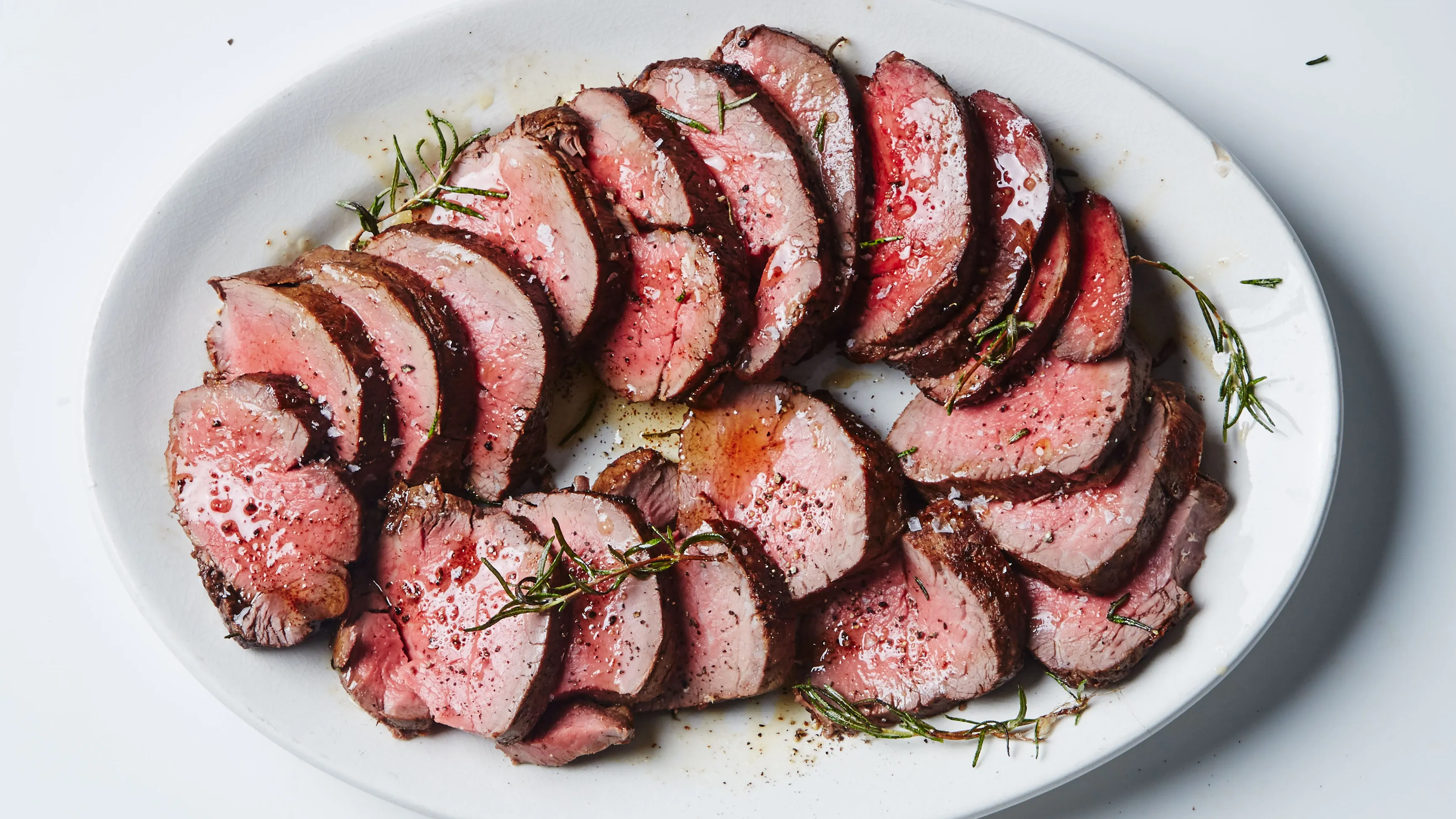Sliced beef tenderloin roast arranged on a platter with rosemary sprigs, flaky salt, and a small bowl of glossy pan sauce on a warm wood table