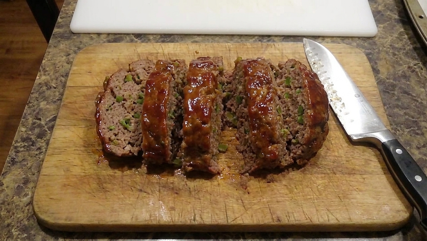Sliced glazed meatloaf on a wooden cutting board with a chef's knife nearby