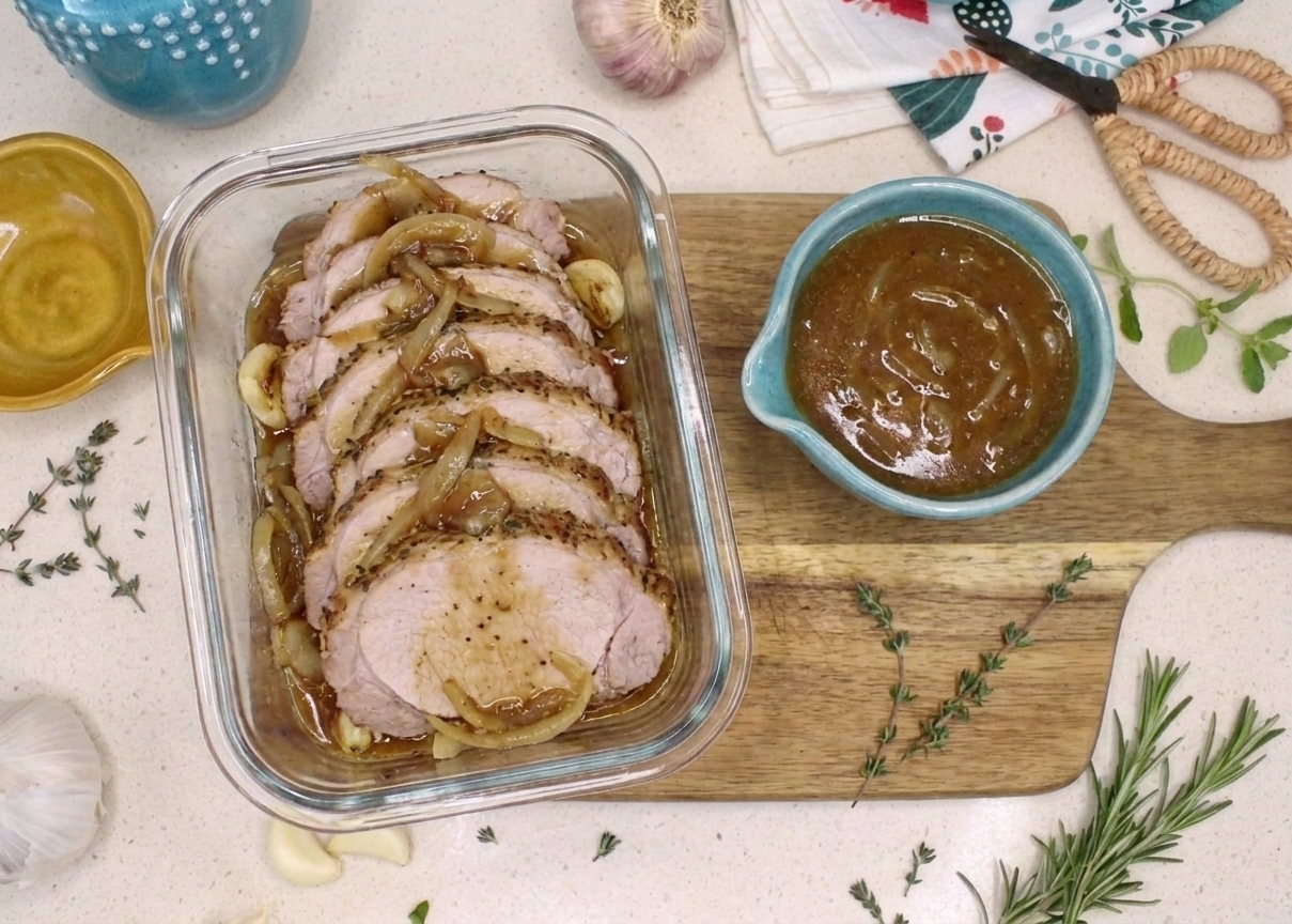 Sliced leftover pork roast arranged in a glass storage container with a small separate container of gravy on a kitchen counter
