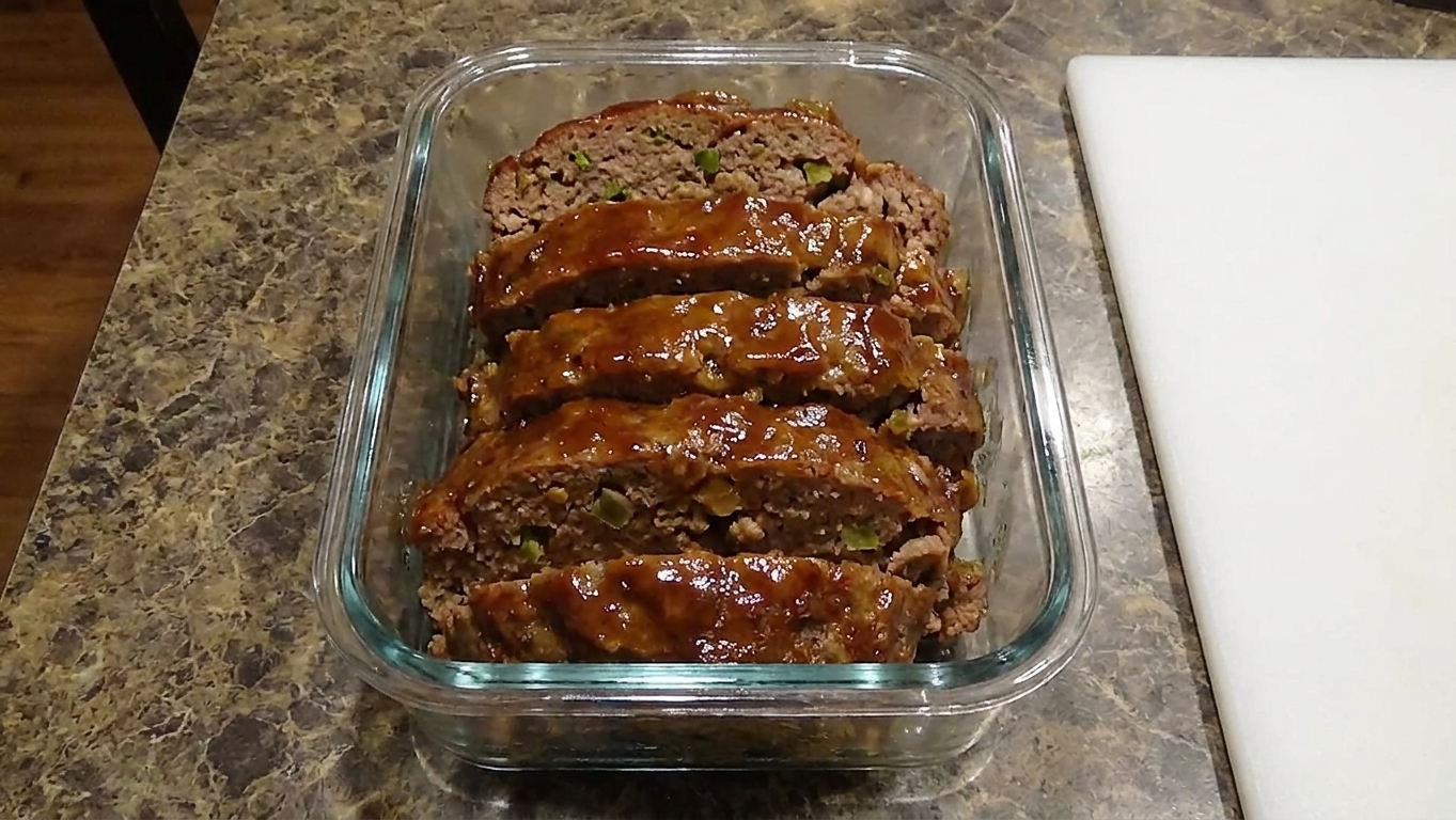 Sliced meatloaf stacked in a glass meal prep container with a bit of glaze visible, set on a kitchen counter