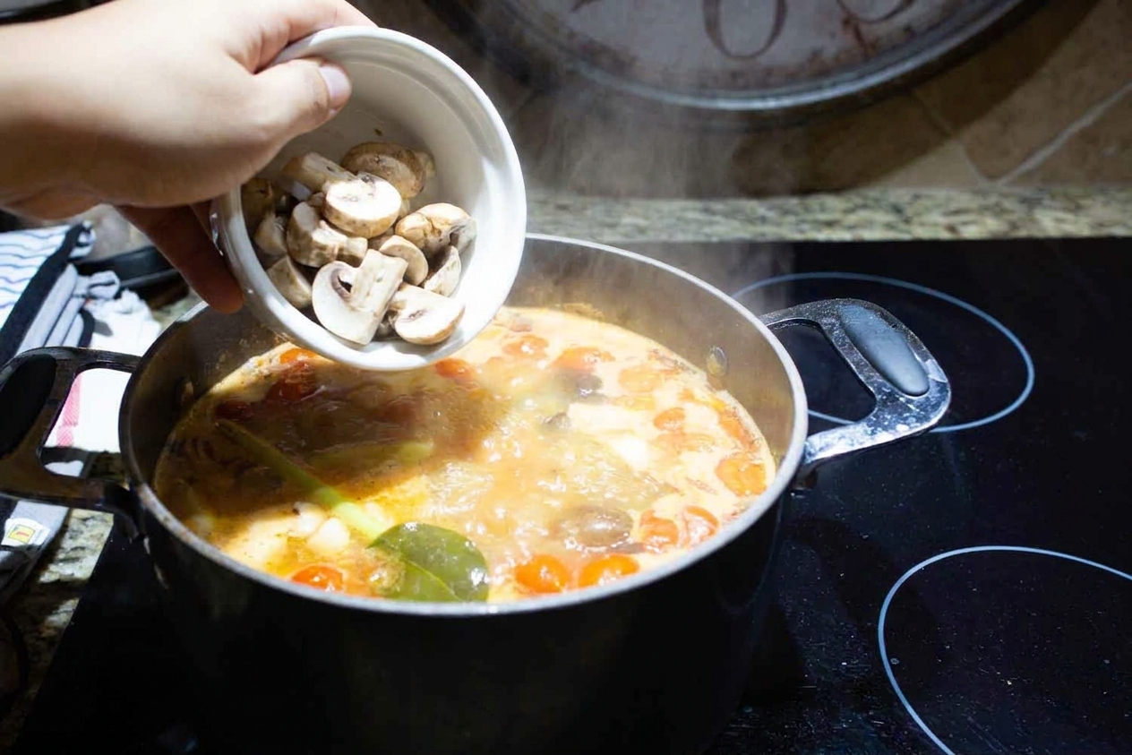Sliced mushrooms being added to a simmering pot of tom yum broth on a stovetop