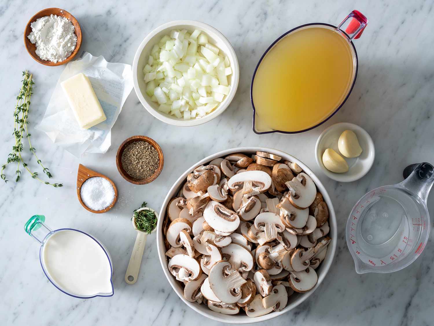 Sliced mushrooms, chopped onion, minced garlic, broth, cream, butter, olive oil, thyme, salt, pepper, and a lemon laid out on a tidy kitchen counter in consistent warm light