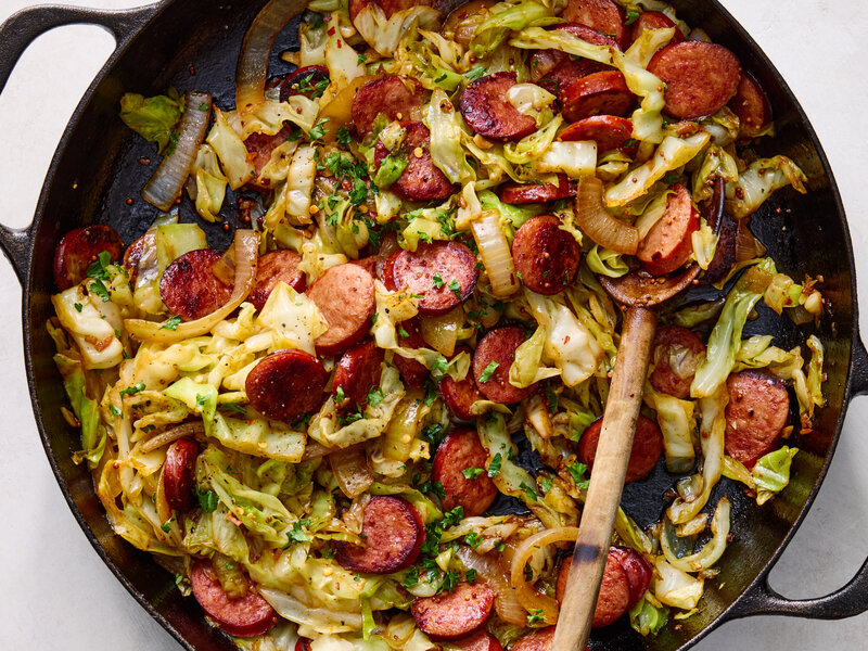 Sliced smoked sausage coins and a pile of thinly sliced green cabbage on a cutting board beside a chef's knife