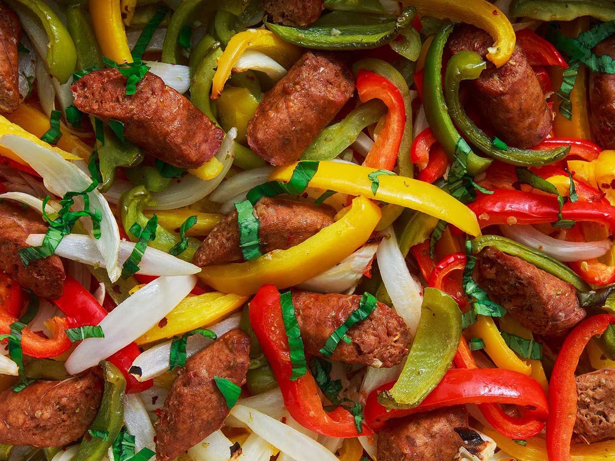 Sliced smoked sausage coins with sliced bell peppers and onions arranged on a cutting board beside a knife on a kitchen counter