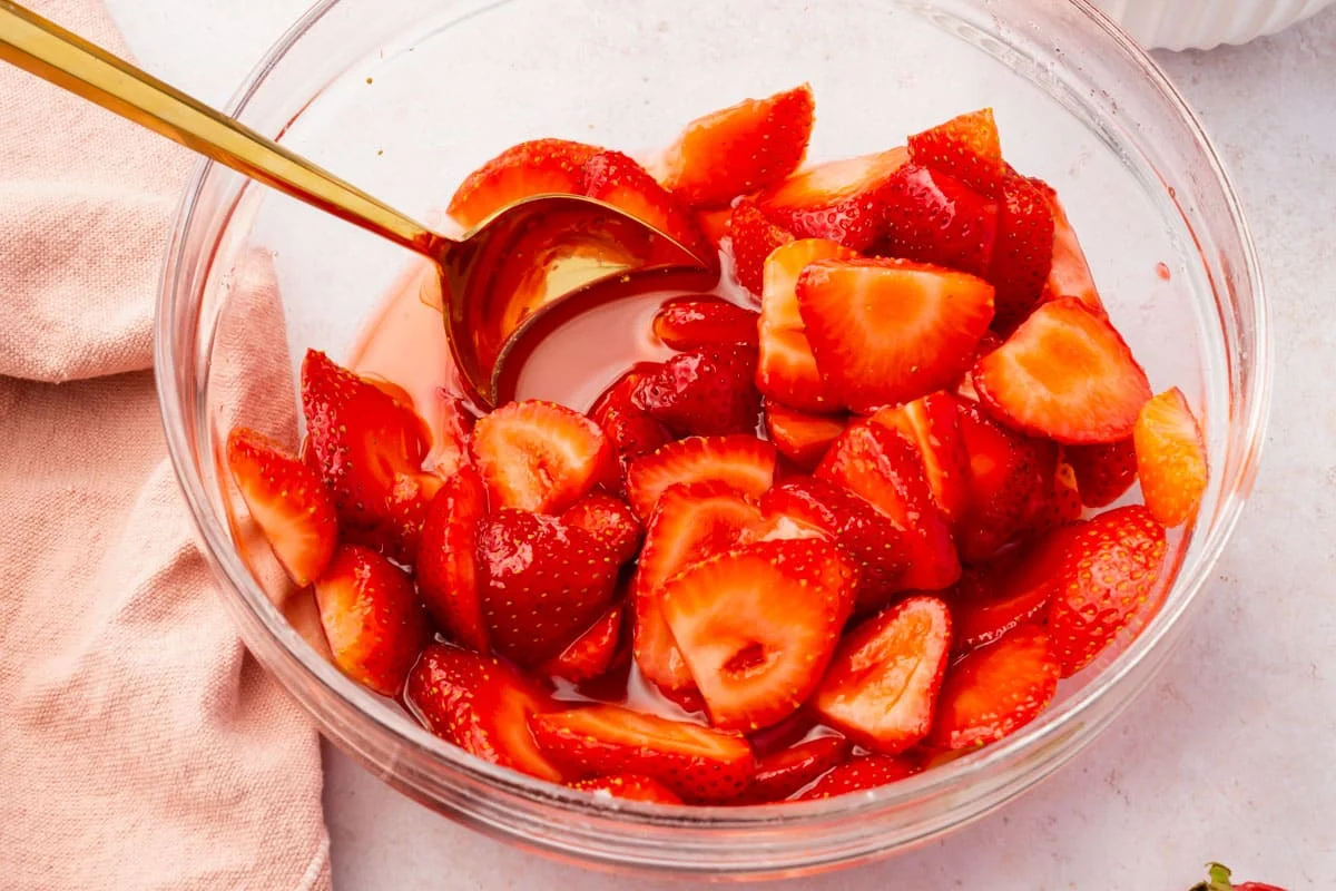 Sliced strawberries in a glass mixing bowl being tossed with sugar and lime juice on a light kitchen counter