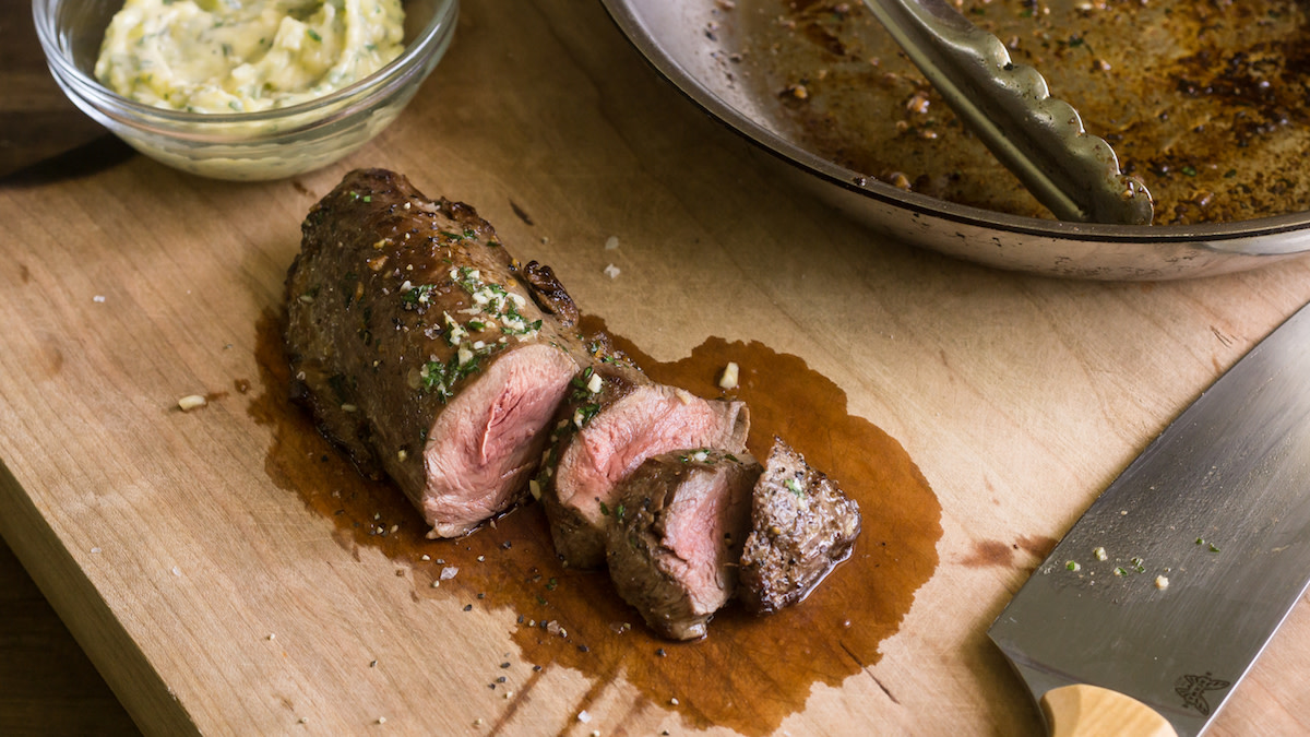 Sliced venison tenderloin fanned on a white plate with melted garlic herb butter, flaky salt, and a few rosemary sprigs on a warm wooden table