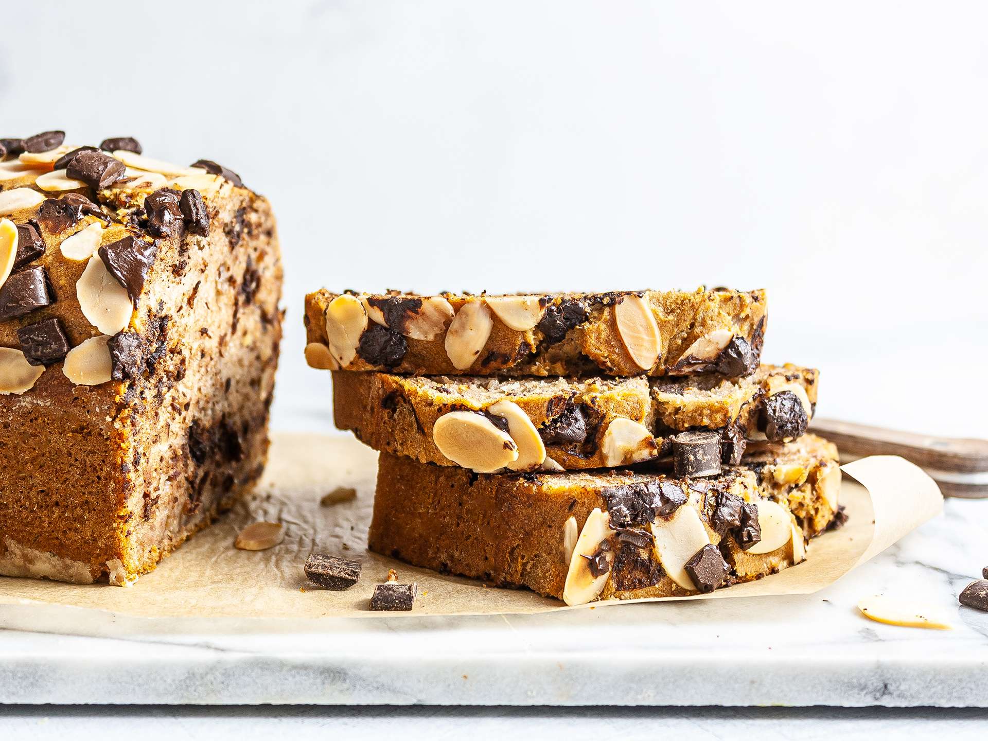 Slices of banana chocolate chip bread stacked in an airtight container on a kitchen counter with a linen towel nearby
