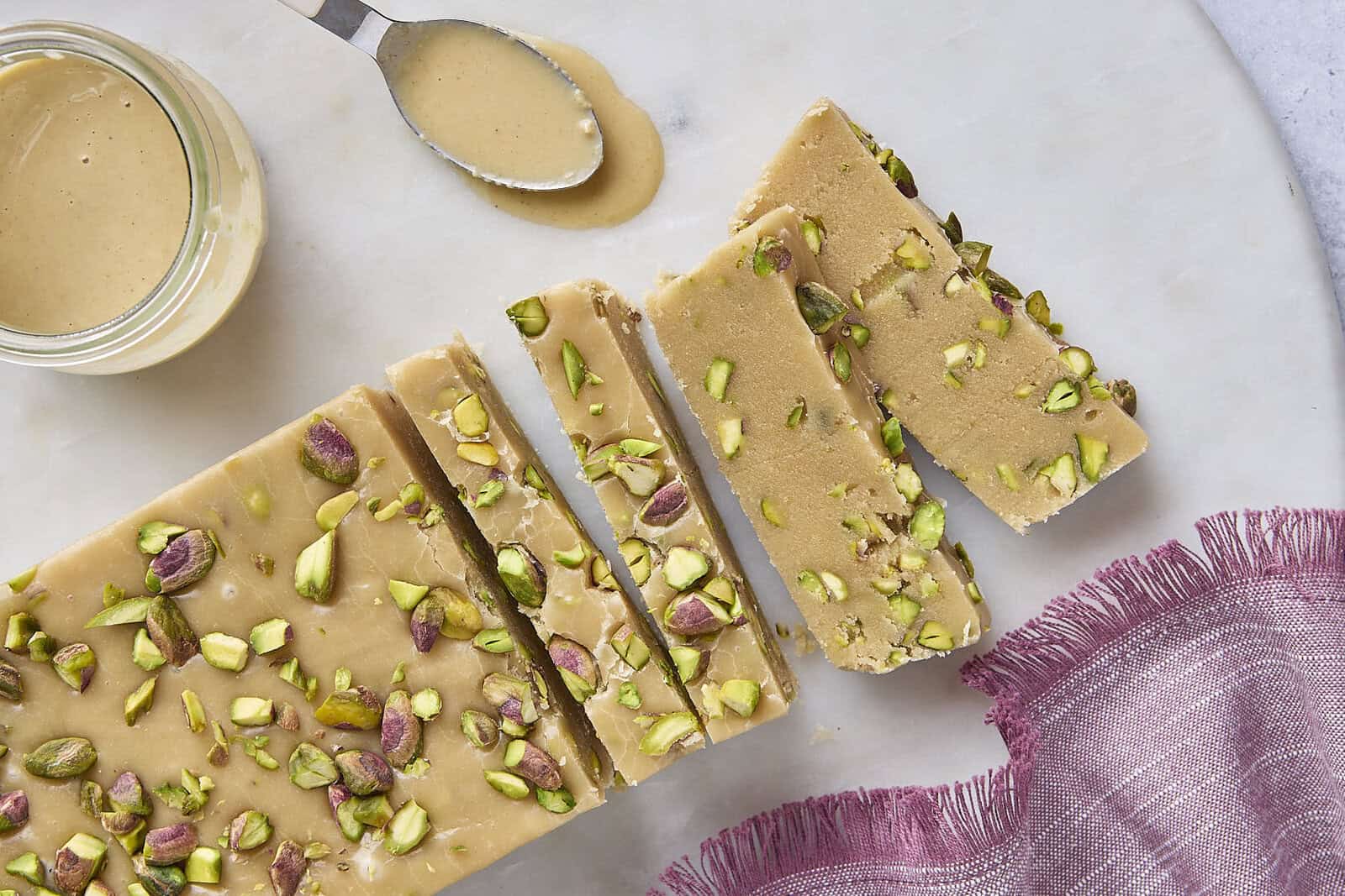 Slices of pistachio halva arranged on a small ceramic platter, showing a pale sesame base with green pistachio pieces, on a warm kitchen table with soft natural light