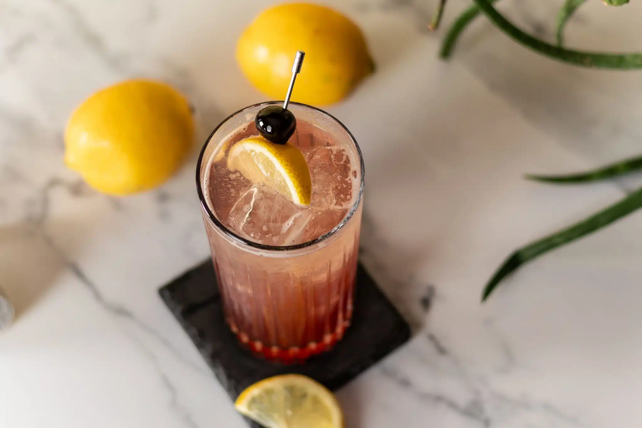 Sloe gin, a fresh lemon, and a small jar of simple syrup arranged on a light kitchen counter beside a jigger and cocktail shaker