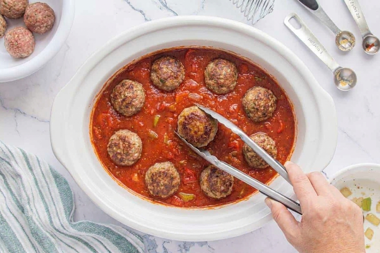 Slow cooker insert filled with red tomato sauce and browned meatballs being nestled in with a spoon