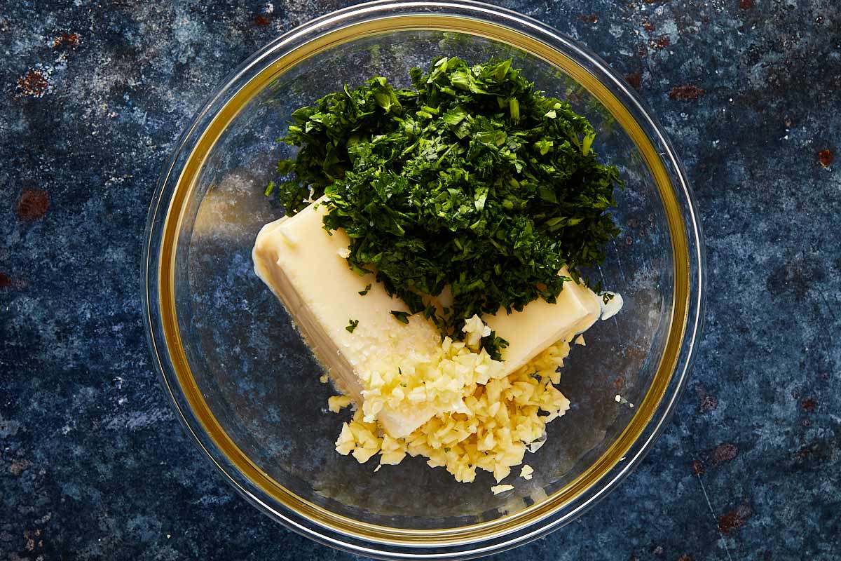 Small bowls holding minced garlic, cubed butter, chopped parsley, thyme, salt, pepper, and lemon halves arranged on a clean kitchen counter