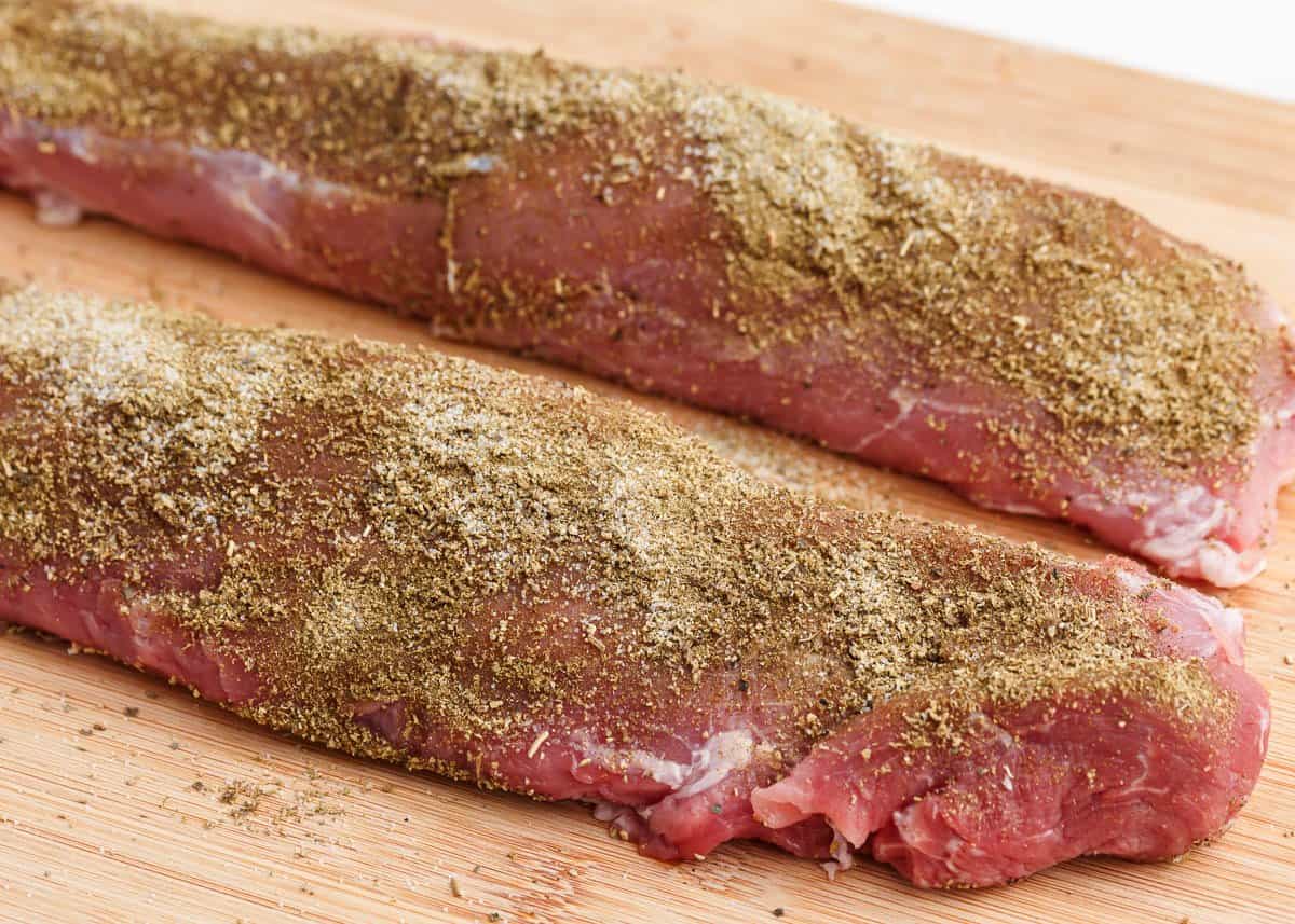 Small bowls of brown sugar, kosher salt, smoked paprika, garlic powder, black pepper, and mustard powder arranged on a kitchen counter next to a raw pork tenderloin