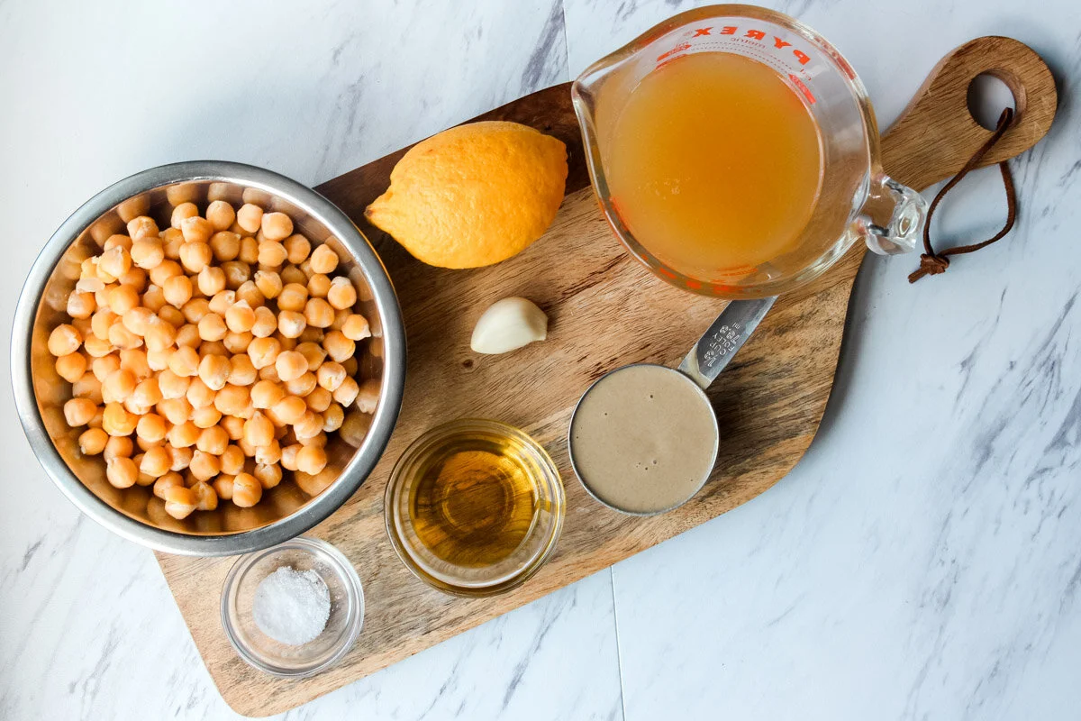 Small bowls of chickpeas, lemon juice, minced garlic, cumin, salt, olive oil, and peanut butter arranged mise en place on a neutral countertop