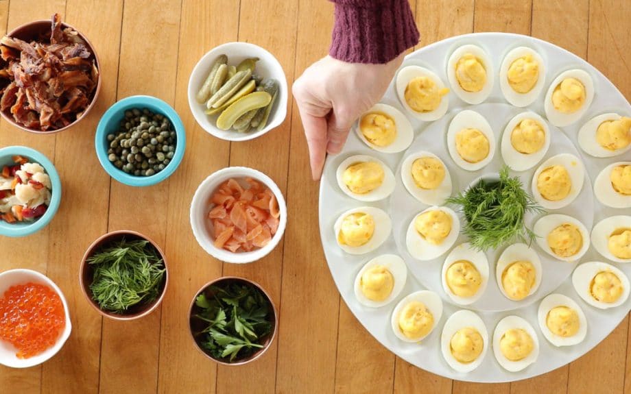 Small bowls of deviled egg toppings on a kitchen counter, including crumbled bacon, chopped pickles, herbs, hot sauce, shredded cheese, and everything bagel seasoning