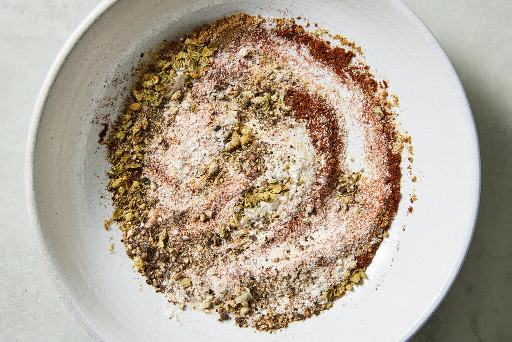 Small bowls of garlic powder, onion powder, oregano, cumin, paprika, black pepper, and salt arranged on a cutting board on a kitchen counter