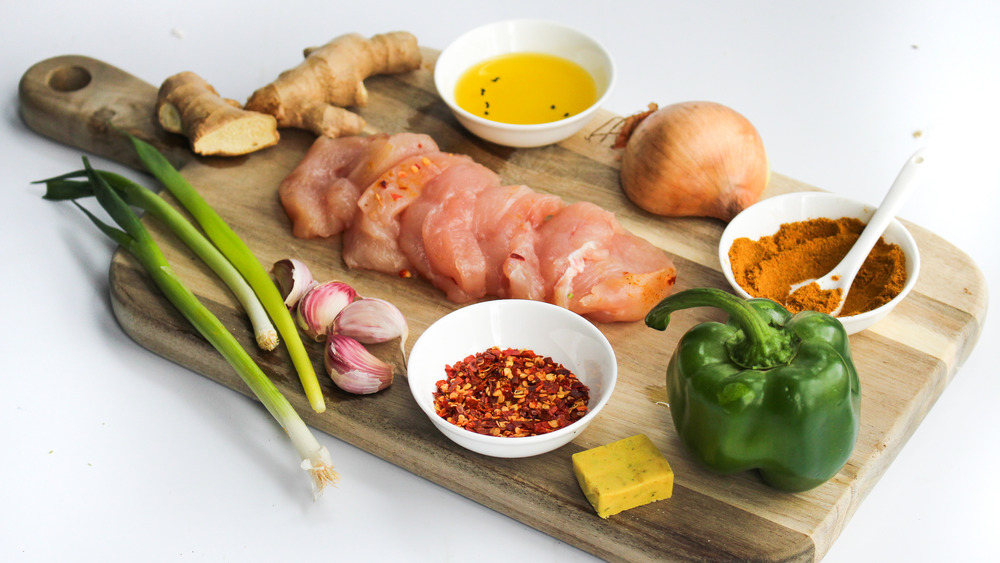 Small bowls of ground cumin, coriander, turmeric, paprika, and garam masala arranged on a wooden cutting board beside fresh ginger and garlic
