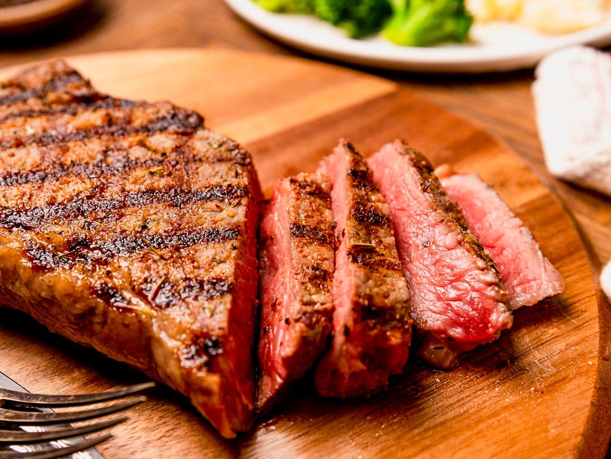 Small bowls of kosher salt, black pepper, garlic, and herbs arranged beside two raw striploin steaks on a wooden cutting board on a bright kitchen counter