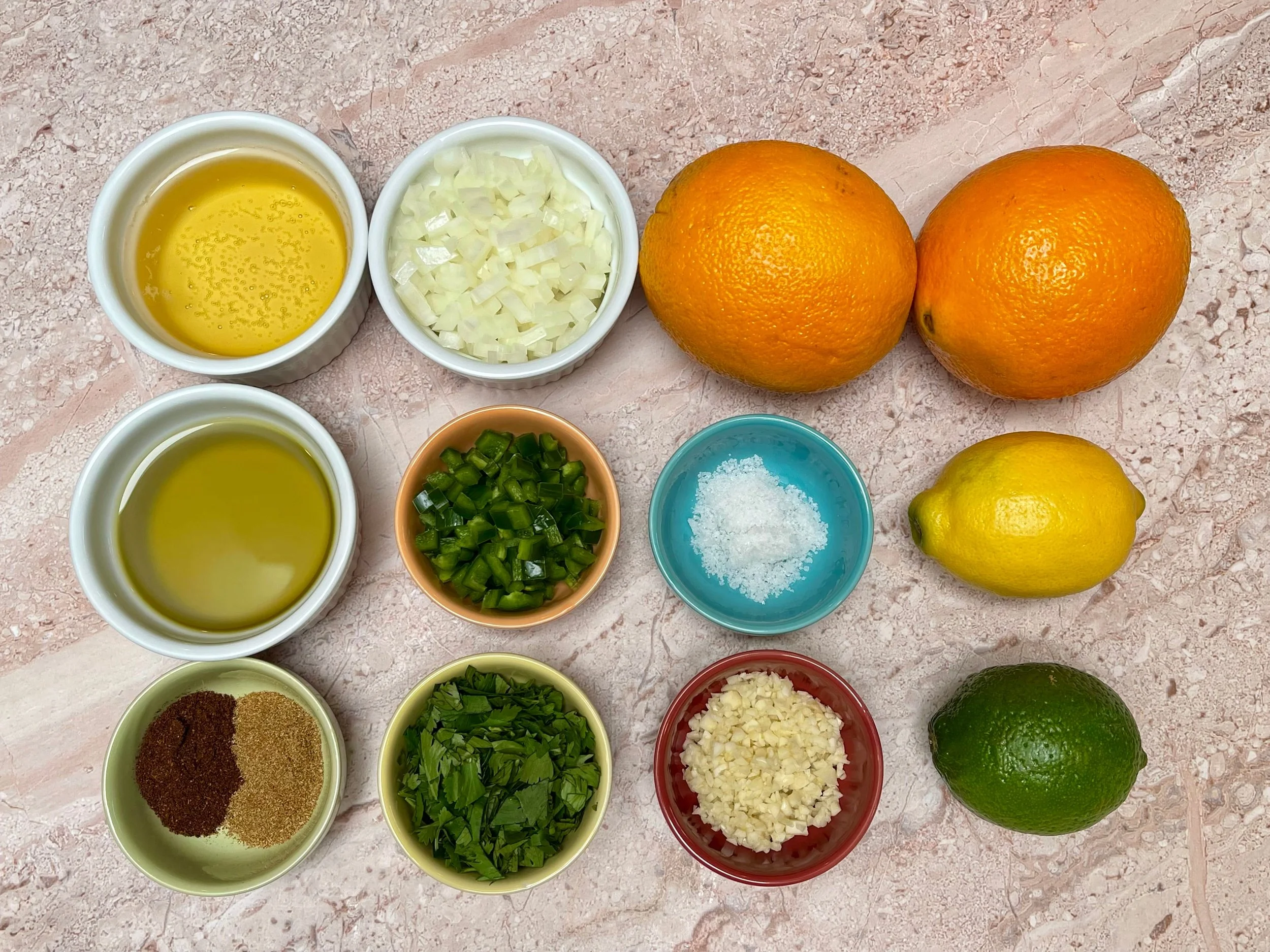 Small bowls of lime juice, orange juice, chopped cilantro, minced garlic, sliced jalapeno, oil, cumin, chili powder, salt, and pepper arranged on a kitchen counter