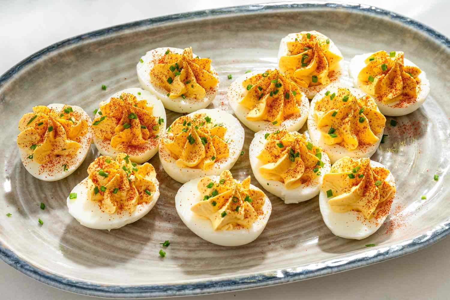 Small bowls of mayonnaise, mustard, vinegar, salt, pepper, and paprika arranged on a light kitchen counter next to whole eggs in their shells