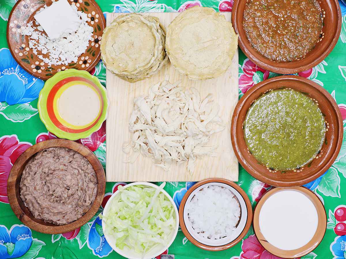 Small bowls of refried beans, shredded chicken, salsa verde, shredded lettuce, queso fresco, crema, and lime wedges arranged on a kitchen counter