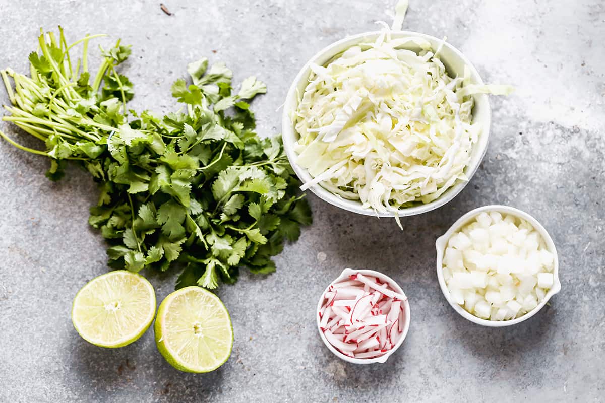 Small bowls of shredded cabbage, sliced radishes, diced onion, chopped cilantro, and lime wedges arranged on a kitchen counter, ready for serving