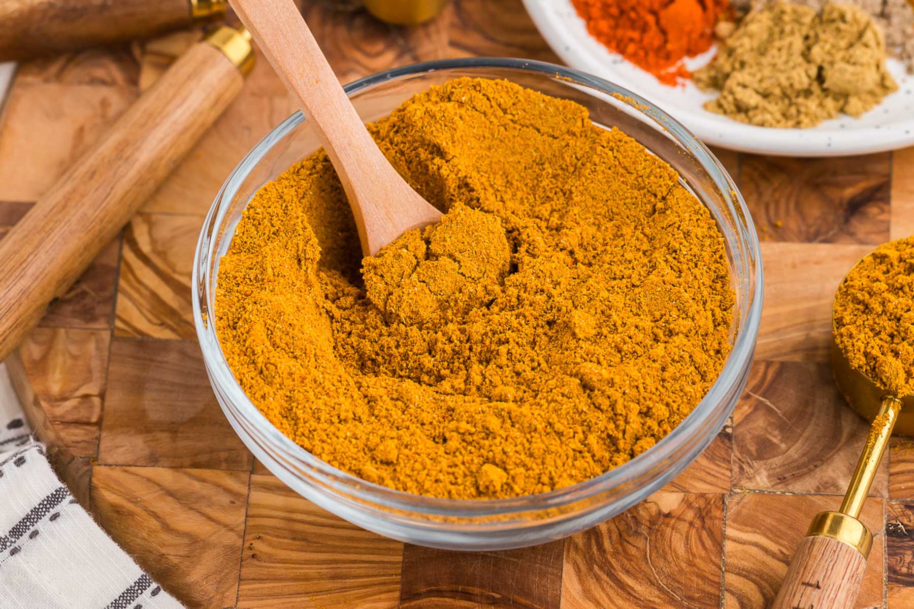 Small bowls of turmeric, cumin, coriander, ginger, paprika, and black pepper arranged on a wooden cutting board in a home kitchen