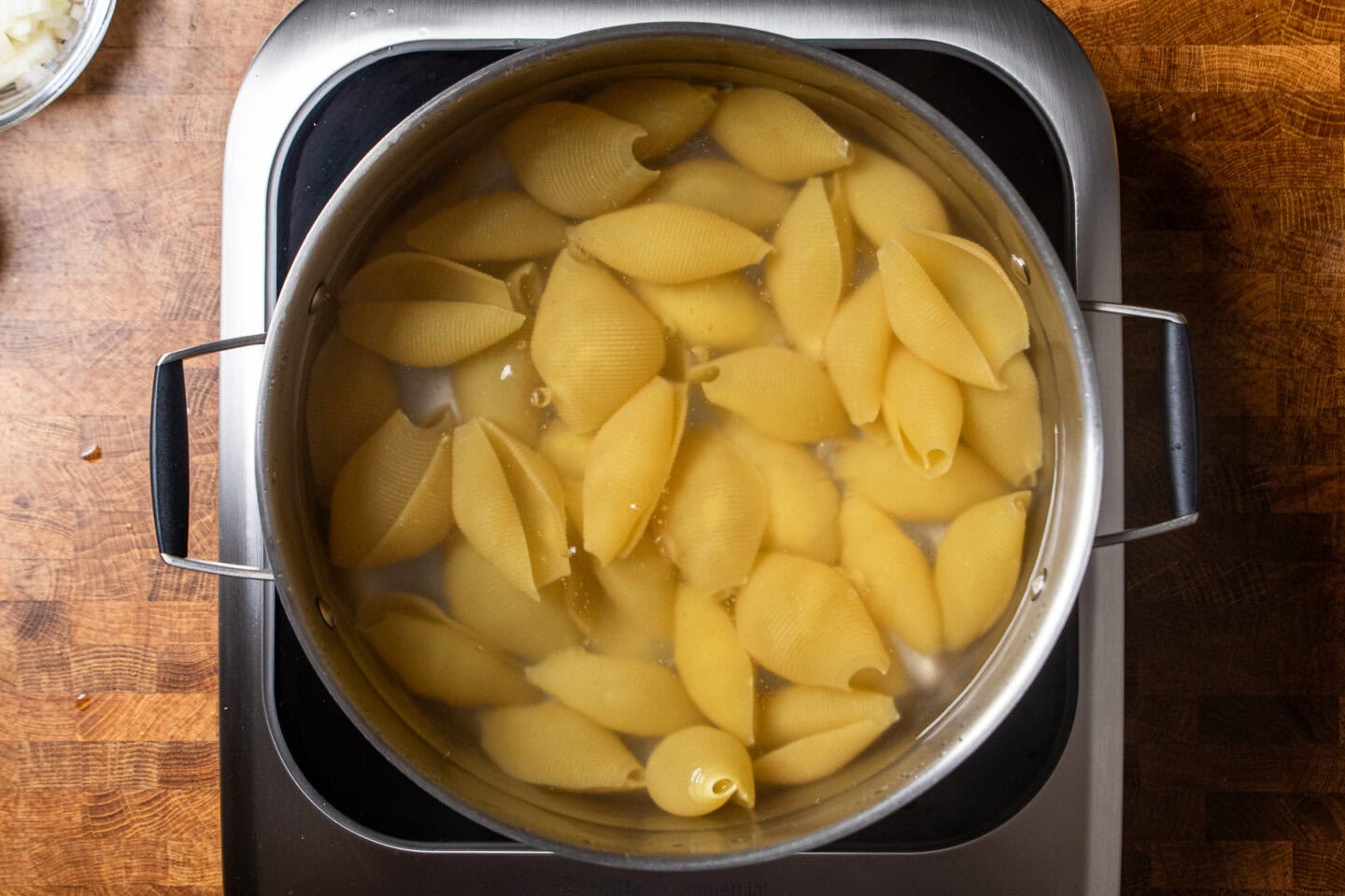 Small pasta shells boiling in a large stainless steel pot on a stovetop with steam rising