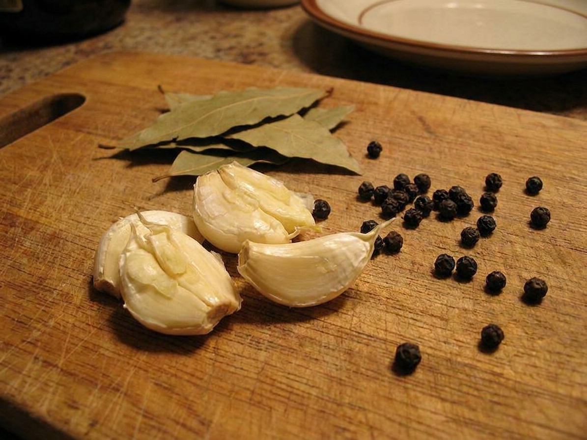 Smashed garlic cloves on a wooden cutting board with bay leaves and whole peppercorns nearby