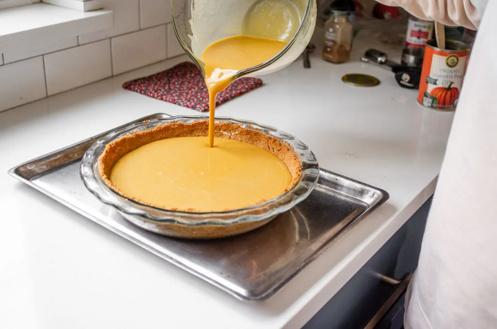 Smooth pumpkin pie filling being poured from a bowl into an unbaked pie crust in a pie dish set on a rimmed baking sheet